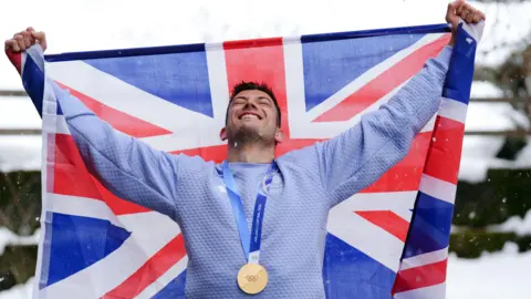 A man wearing a gold Olympic medal is smiling as he looks up to the sky, holding out a large Great Britain flag.