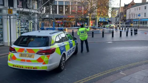 BBC A police officer lifting up tape blocking a city centre street, next to a parked police car