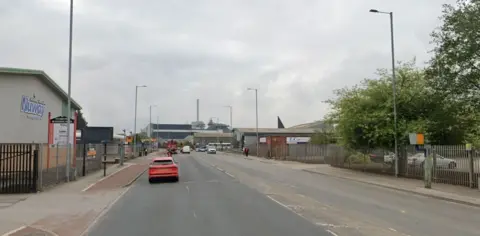 Google A two-lane carriageway with a red car driving southbound. There are speed cameras on both sides of the road and vehicles stopped at red traffic lights in the far distance.
