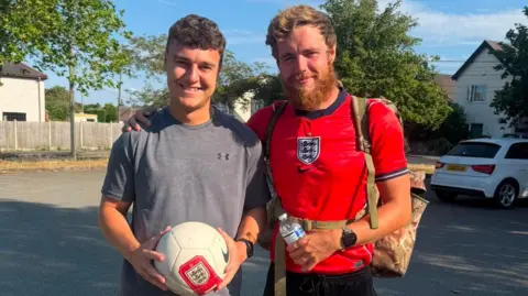James Lewis Two men are standing next to each other smiling, holding a football and water bottle. They are both wearing sports t-shirts and standing in a car park.