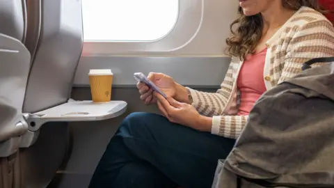 Woman in beige and white striped cardigan, salmon t-shirt and dark blue trousers sitting in a train carriage, looking at mobile phone.