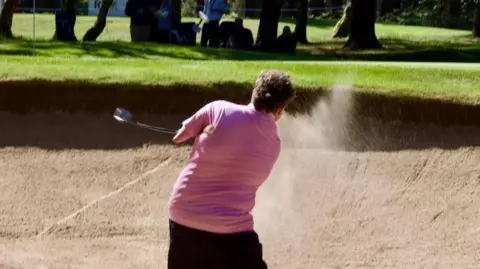 Adam is in a golf bunker hitting the ball out of the sand. He is wearing pink shirt and holding the club under his arms.