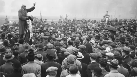 Getty Images 24 September 1933: Berbicara kepada massa di Speakers' Corner di Hyde Park, anggota parlemen Komunis Saklatvala Shapurji menyerukan pembebasan tersangka Kebakaran Reichstag di Jerman. Kebakaran yang membakar gedung parlemen Reichstag diduga dimulai oleh anggota Partai Komunis Marinus van der Lubbe dan memberikan alasan kepada pemerintah Jerman untuk memberlakukan keadaan darurat di seluruh negeri dan menekan penentang rezim Nazi. (Foto oleh Keystone/Getty Images)