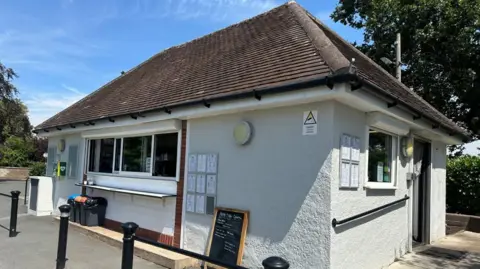 BBC/Charlotte Benton A small white building that is a cafe, it has a rectangular window at the front, that is an ordering hatch and an opening on the side with a chain curtain tied up. The sky is blue in the background and a large tree shadows the back of the building. 