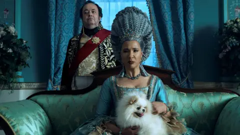 Reuters/Netflix An image from the TV series Bridgeton with Golda Rosheuvel as Queen Charlotte sitting on an opulent green settee with a small white dog on her lap. She is wearing an intricate turquoise silk dress and her blue coloured hair is piled high on her head. 