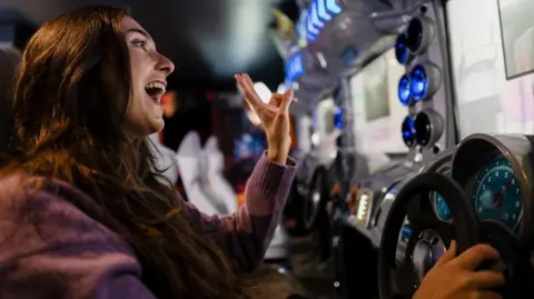 A woman with long brown hair sits an a racing car gaming machine.