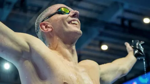 Martin Bennell A smiling man wearing goggles with his arms outstreched after exiting a swimming pool