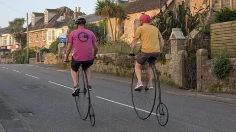 Two riders on penny farthing bicycles cycle along a quiet Cornish street past a stone church.
