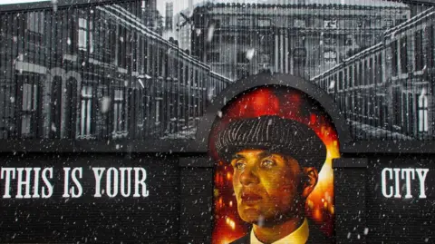 SnipedbyKIM The mural is painted black and says 'this is your city'. The top of the factory wall is filled with painted black and white terraced houses. Thomas Shelby is painted in an archway of the wall.