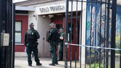 Three PSNI officers standing at the front entrance of Divis Tower. There is blue police tape across a black metal fence. 