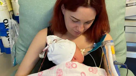 Tia with dark red hair is sitting in a green soft chair wearing a black sleeveless top, looking down at the baby cradled against her chest. The back of Arabella's head, wearing a white hat is visible as is part of the white and pink blanket that covers her. Tubes can be seen coming from her face. Oxygen cannisters are visible in the background.