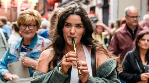 A woman with long brown hair, playing a tin whistle. There is a crowd of people behind her.