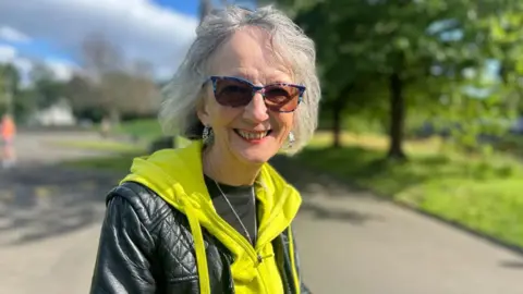Dot Tilbury smiling. She's wearing a black leather jacket and a yellow hooded top. Behind her is a cycleway lined with green trees.