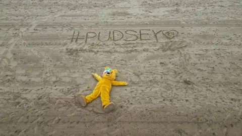 Dale Baxter / BBC Pudsey Bears lies on a beach with his arms outstretched with the Pudsey 24 hashtag written in the sand above his head