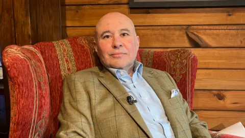 A mid shot of Carl Meerveld sitting in an old-fashioned red armchair. He is bald, and wearing a brown jacket, and light shirt.