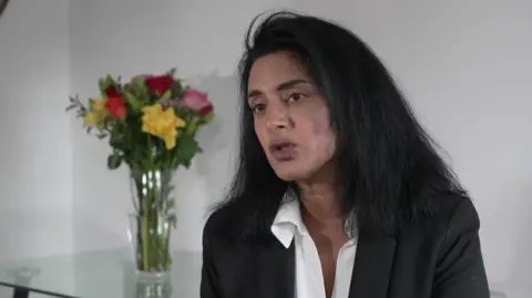 A middle aged woman dressed in a suit jacket and white shirt with long dark hair sits in a neutral setting. There is a vase of flowers on a table behind her. 