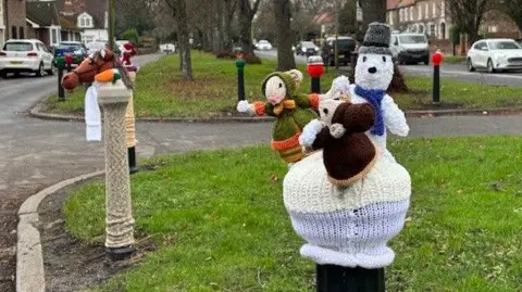 Haughton Ladies Who Knit A wide residential street separated by a central grass verge with a line of  well-established trees. Among the knitted characters sitting on bollards are a horse, Father Christmas and three mice characters in Dickensian dress.