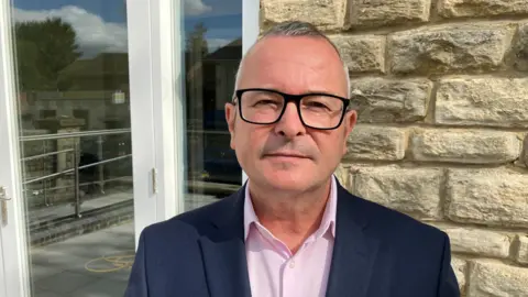 Lee Barron in a pink shirt and navy blazer with glasses stands in front of a sand-coloured brick wall, looking at the camera. 