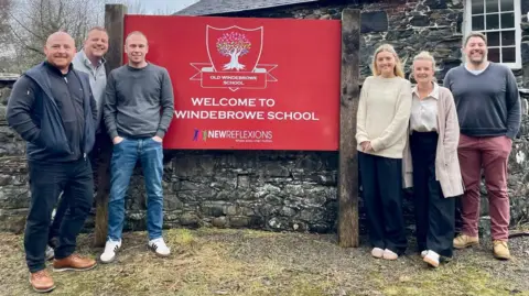 Six people stand on either side of a school sign and are smiling. The red sign, which is partially obscured by a person standing in front of it, reads: "Welcome to... Windebrowe School". The school building behind them is made of grey stone.