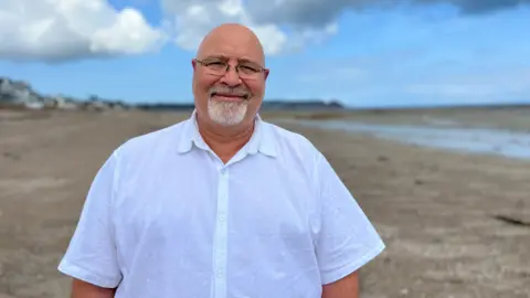 Kenan has a beard and is wearing glasses and a white short-sleeved shirt stands on a wide, mostly empty beach under a cloudy sky. Buildings are visible in the distance to the left, and the ocean stretches out to the right.