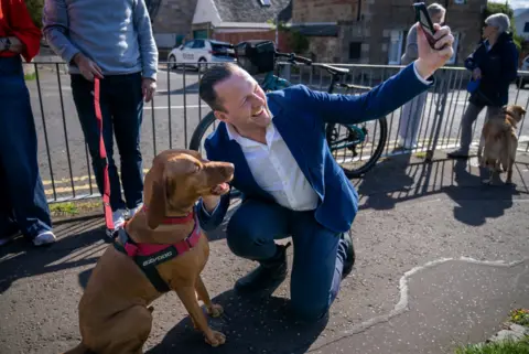 PA Media Man in a suit crouching on a pavement taking a selfie with a brown dog on a lead, while other people and dogs stand nearby on a residential street
