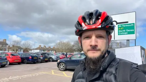 Vikki Irwin/BBC Alexander Gross is wearing a black and red cycle helmet and has a beard. He is wearing a black puffercoat. He is standing by a car park.