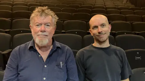 Jon Wright/BBC Rob John and Merlyn Haycroft sit on chairs in a theatre
