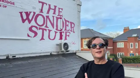 Miles is stood near a white building with his band's name painted on it in pink paint. He is wearing a black top and sunglasses and is posing with his arms folded.