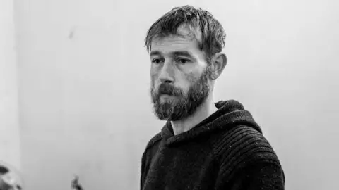Black and white image of Chris Garrett looking into the distance. He is wearing a dark coloured heavy hooded jumper