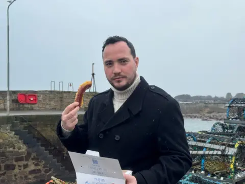 A man in a dark coat and cream turtleneck stands on a harbour pier on a grey day, holding a battered sausage in one hand and an open fish and chips box in the other. Lobster pots and stone harbour walls are visible behind him, with the sea in the background.