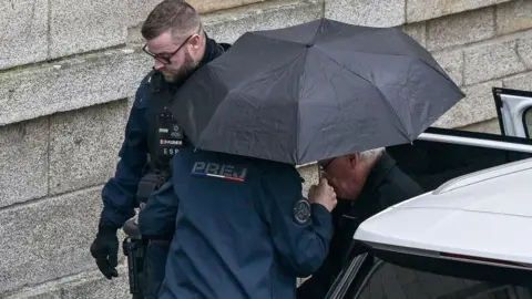 DAMIEN MEYER/AFP A man exits a car helped by police, one of whom is holding an umbrella