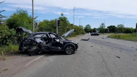 Crown Prosecution Service The black car is shown badly damaged on the side of a road, it is crumpled with both the boot and bonnet up and multiple smashed windows as well as damage to the body of the car.