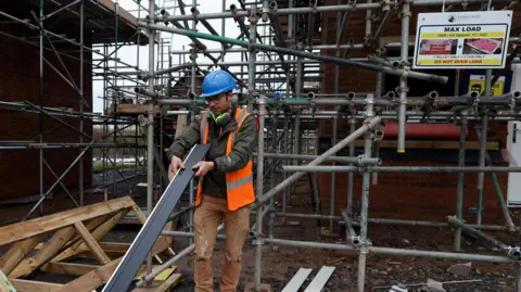 REUTERS/Temilade Adelaja Construction worker in a blue hard hat at house building site, with metal scaffolding in the background and a warning signe. The builder is holding a long piece of plastic.