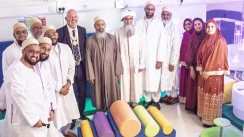 A group of people are standing in a curved line looking at the camera. There is a group of men and women, some are wearing bright colours, others are in white. They are standing in the sensory room. 
