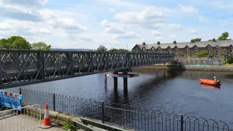 Leeds City Council A footbridge running from one side of a river to another. A man in an orange kayak is on the water approaching it. Some houses can be seen on the other side of the river.