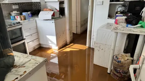 Muddy water remains on the floor while a tide mark can clearly be seen on the walls and more than half way up the side of a white washing machine and grey oven in this kitchen
