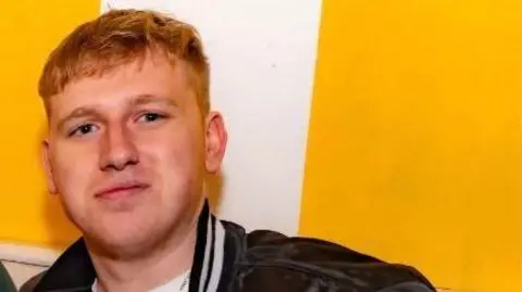 Jake Davies A man with short ginger hair smiling and looking at the camera. He is wearing a black jacket and white top. He is standing in front of a white and yellow wall.