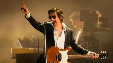 Getty Images Alex Turner singing on stage. He is wearing sunglasses and pointing towards the sky.