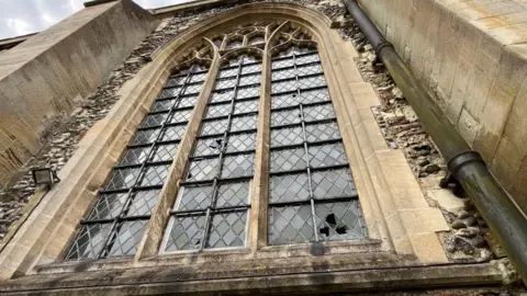 Andrew Turner/BBC One of the large north-facing windows on St Nicholas Minster; a trio of gothic arched window apertures, with diamond leaded glass set within, featuring a rectangular border. The window is made of sandstone and set within a flint wall.