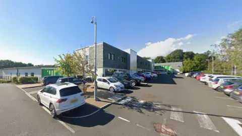 A street view image of Ferndene which is a modern, rectangular building with grey bricks on the front of the building. There are cars parked in front. Large trees can be seen behind the building.