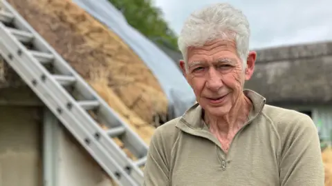 Alan Lewis is smiling at the camera. He has grey hair and is wearing a beige 3/4 zip jumper. Behind him is a ladder and the edge of a thatched roof. 