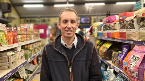 Paul Moseley/BBC Roy Hagan is wearing a fleece-type cardigan on top of a dark jumper and shirt. He is standing in a shop aisle. The aisle's shelves are lined with dog food products.