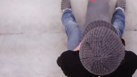 A general view of a boy, who is sat down and is pictured from above. He is wearing a grey flat cap, wearing a black T-shirt and blue jeans. 