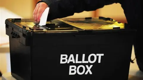 A man's hand putting a folded voting paper into a slit in a lid of a large black box with the words "Ballot Box" in white capital letters on the side.