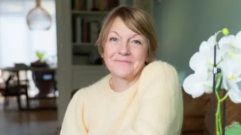 Annie sitting in a furnished room with a white flower in the foreground. She has short brown hair and is wearing a yellow jumper.