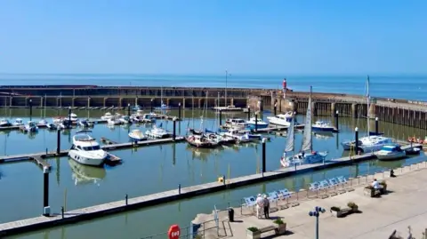 Rob Kean Lots of boats floating in Watchet Harbour. It is a bright sunny day and the blue sky is reflected in the still mirrored water.