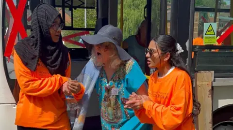 A distressed looking woman wearing a large black sun hat with a big rim is being helped off a bus by two younger people wearing bright orange tops. Some of the bus windows have large red "X" shapes on them. Trees and a red brick building are behind the bus.