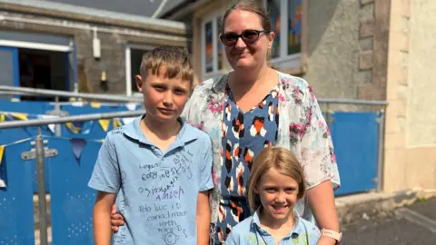 A woman with her arms around two young children who are wearing light blue polo shirts which have been signed
