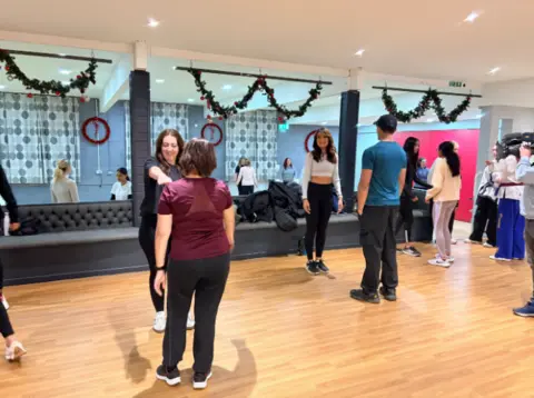 Women and one man in gym wear stand opposite one another at the class as they learn techniques. Christmas decorations can be seen hanging from the ceiling in the background.