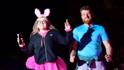 Nick Butcher Photography/St Elizabeth Hospice A woman wearing pink bunny ears, a pink tutu, and a black coat raises her arms in the air and smiles. She holds her phone in one hand. A man next to her is wearing a blue T-shirt. He is also smiling.